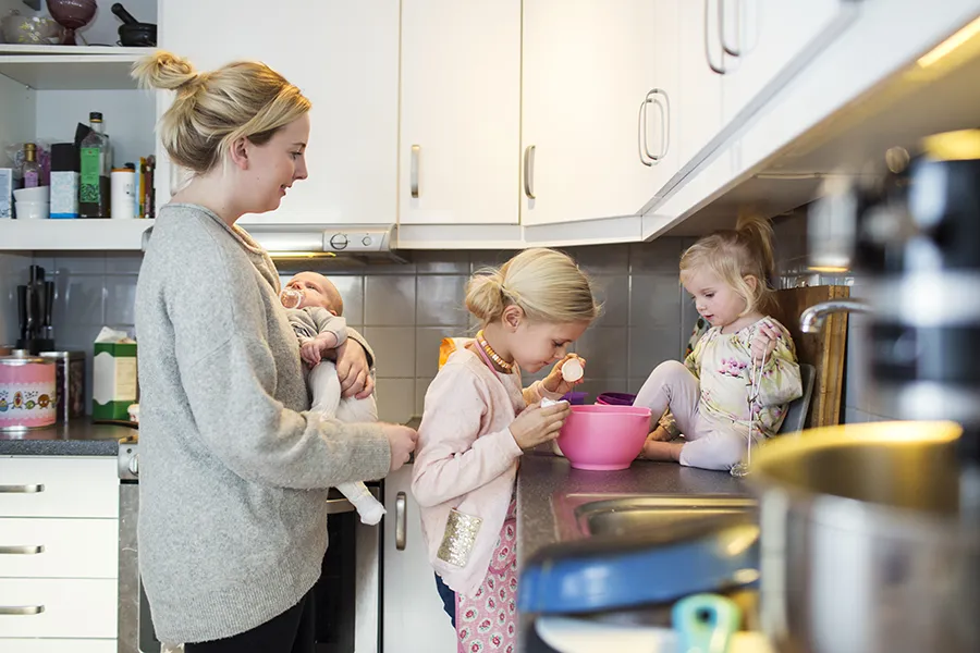 Mother holding her baby while two other children make panacakes in kichen of her rented property.