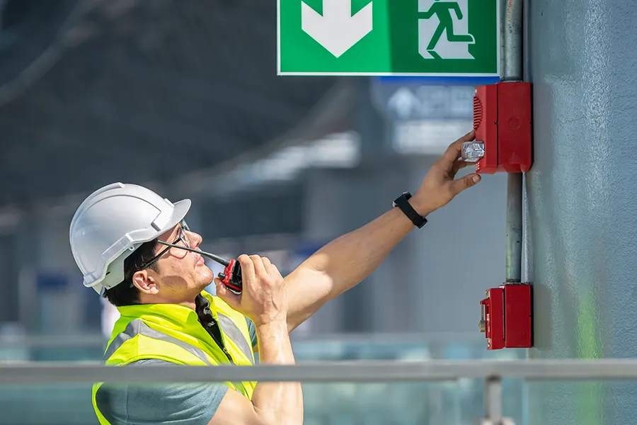 Fire Safety Regulations A fire safety engineer wearing a hi-visibility vest and a hard hat, checking the fire alarm system in a large, open plan building, whilst talking on a walkie-talkie.