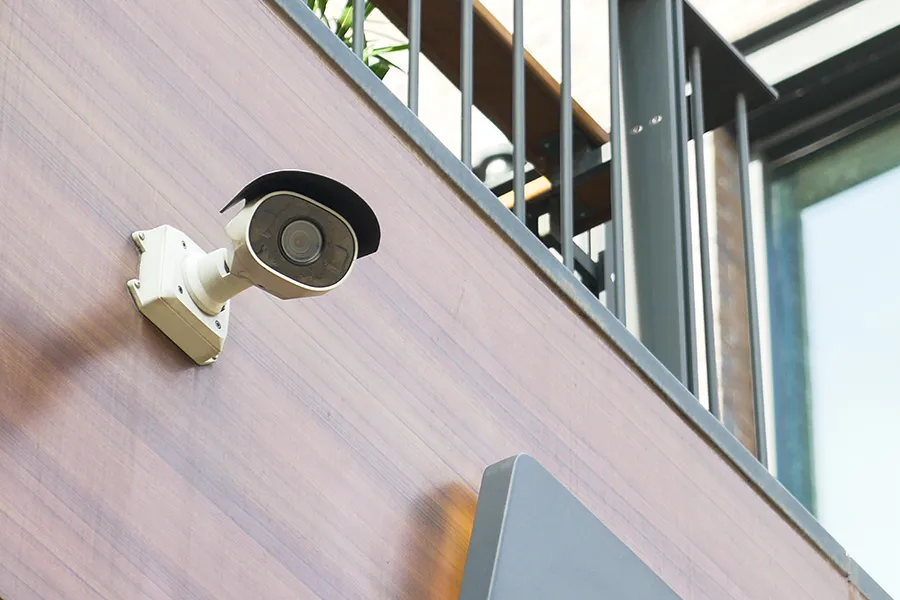 Part of a home CCTV installation, a security camera is mounted high up on the side of a wall with terrace above.