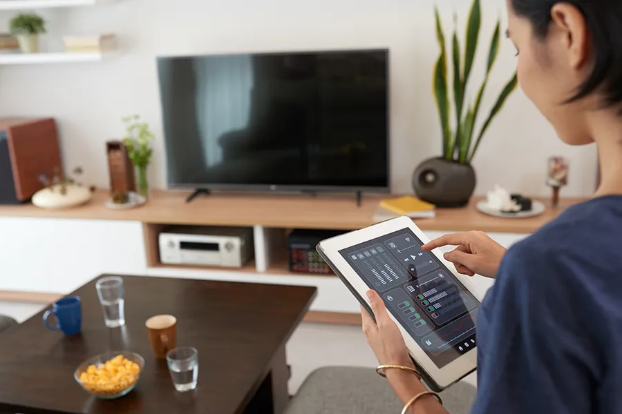 A woman in her living room, holding a tablet that is controlling her smart home entertainment system.