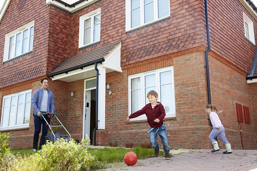 Children playing in the front garden as their father mows the lawn outside their rental property.