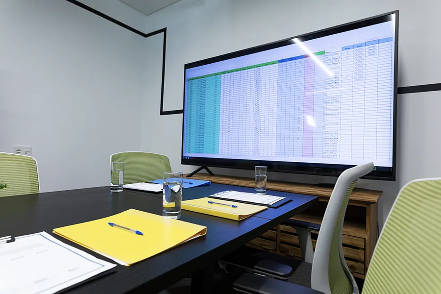A meeting room with a large screen, that has no visible cables, showing a speadsheet and a meeting table with documents and glasses of water.